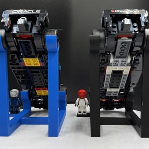 May include: Two black toy race cars with the Oracle and MoneyGram logos are displayed on stands. One stand is blue, the other is black. Small toy figures stand in front of each car. The cars are made of black blocks.