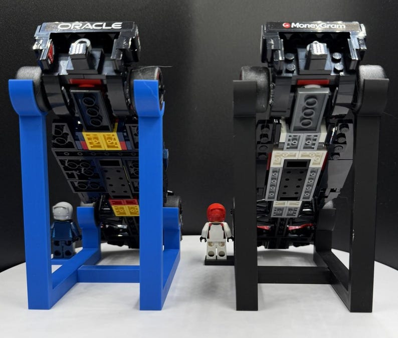 May include: Two black toy race cars with the Oracle and MoneyGram logos are displayed on stands. One stand is blue, the other is black. Small toy figures stand in front of each car. The cars are made of black blocks.