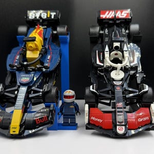 May include: Two Formula 1 race cars, one navy blue and yellow, the other black and red, with matching driver figurines. The navy car has "BYBIT" and "Red Bull" branding. The black car has "Haas" and "Money Gram" branding.