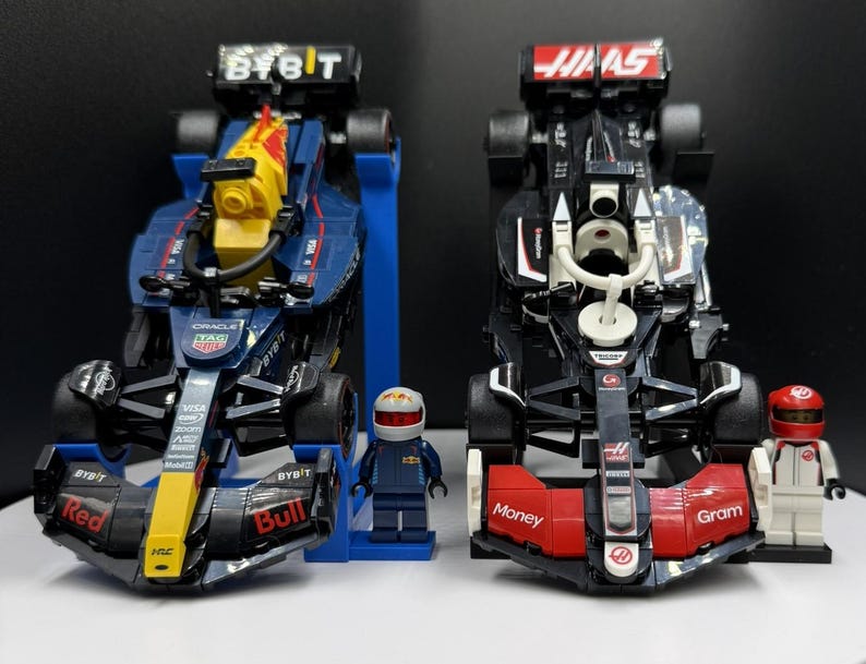 May include: Two Formula 1 race cars, one navy blue and yellow, the other black and red, with matching driver figurines. The navy car has "BYBIT" and "Red Bull" branding. The black car has "Haas" and "Money Gram" branding.