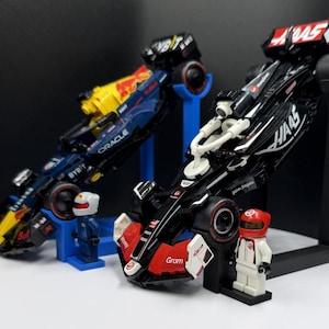 May include: Two toy Formula 1 race cars, one black and red, the other blue and yellow, are displayed on stands. Each car has sponsor logos and is accompanied by a small figure in a racing suit and helmet. The cars are angled, suggesting speed.
