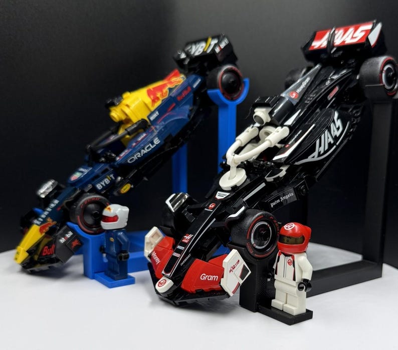 May include: Two toy Formula 1 race cars, one black and red, the other blue and yellow, are displayed on stands. Each car has sponsor logos and is accompanied by a small figure in a racing suit and helmet. The cars are angled, suggesting speed.