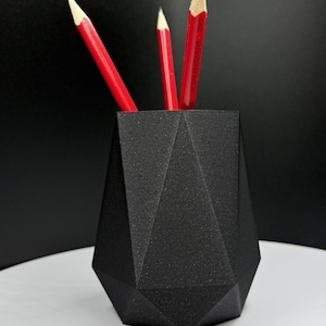 Può includere: Un portapenne geometrico nero con tre matite rosse. Il supporto ha un design sfaccettato e una superficie strutturata. Le matite sono appuntite, rendendolo un elegante accessorio da scrivania.