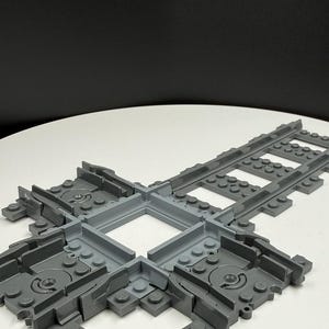 Könnte beinhalten: Ein graues Kunststoff-Spielzeug-Gleisstück. Die Strecke hat eine quadratische Mitte und mehrere Anschlussgleise. Das Teil ist so konzipiert, dass Züge die Richtung wechseln können. Die Strecke besteht aus ineinandergreifenden Kunststoffbausteinen.