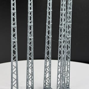 May include: A collection of gray, miniature lattice towers, likely for a model or diorama. Each tower has a square base and a tall, open-frame structure with cross-bracing. The towers are arranged in a row against a dark background.