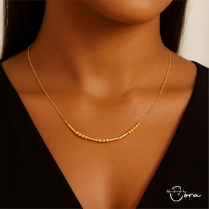 May include: A delicate gold necklace with small beads and cylindrical bars. The necklace is shown against a dark background, highlighting its intricate design. The jewelry is a simple yet elegant accessory.