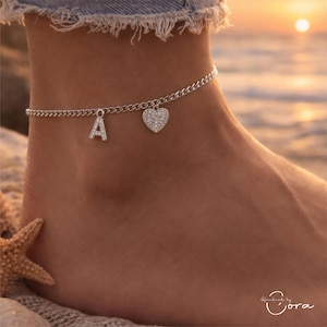 May include: A silver anklet with a chain design. The anklet features two charms: a letter "A" and a heart, both adorned with small, sparkling stones. The anklet is worn on an ankle, with a beach background.
