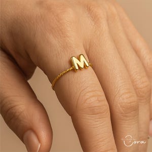 May include: A gold-colored ring featuring the letter "M" as the centerpiece. The ring has a twisted band design. The ring is worn on a finger.