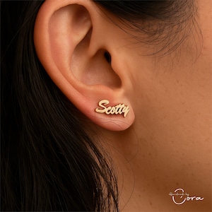 Può includere: Orecchini a lobo color oro con il nome "Scotty" scritto in corsivo. Gli orecchini sono indossati su un orecchio, mostrando il design. Lo sfondo è un primo piano della pelle e dei capelli.