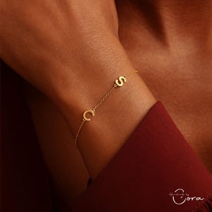 May include: A delicate gold bracelet featuring the letters "C" and "S" connected by a fine chain. The bracelet is worn on a wrist, with a red garment in the background. The bracelet is a personalized jewellery item.
