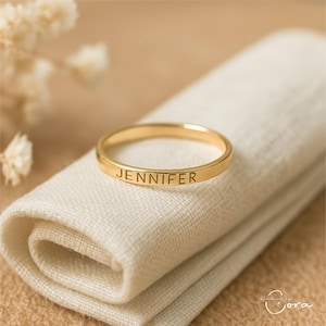 May include: A gold ring engraved with the name "JENNIFER". The ring sits on a folded, off-white cloth. The background is a soft, neutral colour, with small, white flowers. The ring is a simple band design.