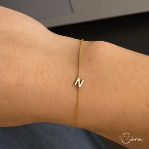 May include: A gold chain bracelet featuring the letter "N" pendant. The bracelet is worn on a wrist. The background is blurred, showing a person's arm and a dark surface. The bracelet is a delicate piece of jewellery.