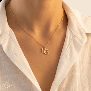 Op de afbeelding: Een goudkleurige ketting met een fijne schakelketting en een hanger bestaande uit de in elkaar verstrengelde hoofdletters D en G. De hanger hangt centraal aan de ketting, die op het sleutelbeen rust en een gepolijste afwerking heeft.