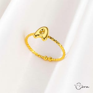 Puede incluir: Anillo dorado con la letra "A" en un diseño llamativo. La banda del anillo presenta un diseño trenzado con pequeños detalles esféricos. El anillo se presenta sobre un fondo blanco.