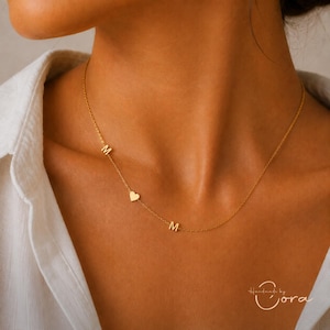 Op de afbeelding: Een goudkleurige ketting met een fijne schakelketting voorzien van twee letter M-bedels en een klein hartvormig bedeltje die asymmetrisch langs de ketting zijn geplaatst. De ketting rust op de hals en heeft een gepolijste metalen afwerking.