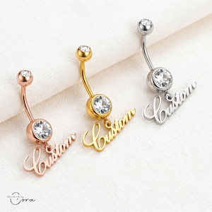 May include: Three belly button rings in rose gold, gold, and silver. Each ring has a curved barbell with a clear gem at the top and bottom. A cursive "Custom" charm hangs from the bottom gem. The background is a white fabric.