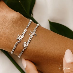 Puede incluir: Dos pulseras de plata con nombres, una con "Kiara" y otra con "Brooklyn", se muestran en una muñeca. Las pulseras tienen un diseño de cadena de bordillo. Los nombres están en una fuente cursiva.