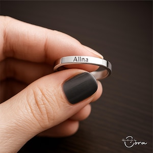 Könnte beinhalten: Ein silberner Ring mit dem eingravierten Namen "Alina". Der Ring wird von einer Person mit dunklem Nagellack gehalten. Der Ring hat ein schlichtes Banddesign.
