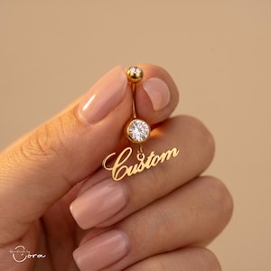 Pode incluir: Um piercing de umbigo dourado com uma pedra preciosa redonda e transparente e a palavra "Custom" em fonte cursiva. O anel tem uma pequena pedra preciosa redonda no topo. O fundo é de cor neutra.