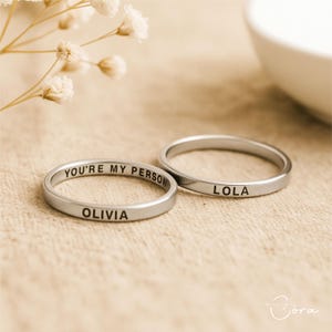 May include: Two silver rings with engraved text. One ring reads "YOU'RE MY PERSON" and "OLIVIA". The other ring reads "LOLA". The rings are on a beige surface, with white flowers in the background.