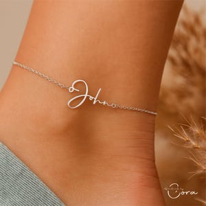Può includere: Una delicata cavigliera d'argento con il nome "John" in corsivo. La cavigliera è indossata su una caviglia, mettendo in mostra il gioiello personalizzato. La catena è sottile e il nome è elegantemente stilizzato.