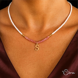 May include: A delicate necklace featuring a white beaded chain that transitions to pink gemstone beads. A gold-colored pendant in the shape of the letter "S" hangs from the center. The necklace is worn around the neck.