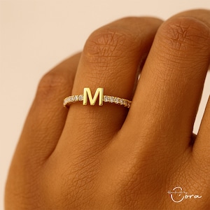 May include: A gold-colored ring with the letter "M" in the center. The band is encrusted with small, clear gemstones. The ring is on a finger, showcasing the jewelry's design and sparkle. A delicate piece of personalized jewelry.