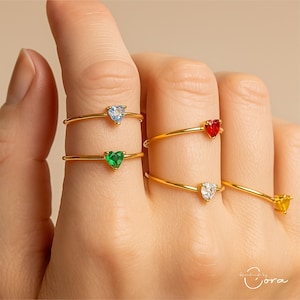 May include: A selection of gold rings, each with a heart-shaped gemstone in shades of light blue, green, red, white, and yellow. The rings have a delicate, minimalist design.