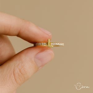 Könnte beinhalten: Ein goldfarbener Ring mit dem Buchstaben "L" und kleinen, klaren Steinen. Der Ring wird zwischen zwei Fingern vor einem neutralen Hintergrund gehalten. Das Band des Rings ist mit kleinen, funkelnden Steinen besetzt.