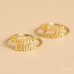 Op de afbeelding: Twee goudkleurige ringen met aangepaste namen. Eén ring spelt "Kaylee Leah" in cursief schrift, de andere "Maya Sarah". De ringen staan tegen een neutrale achtergrond.
