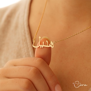 May include: A gold-colored necklace with a delicate chain. The pendant features a name written in Arabic script. The necklace is displayed on a person's neck and finger, showcasing the jewelry's design and style.