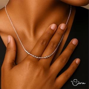 May include: A delicate silver necklace with small, round beads and slender bars. The necklace is shown against a dark background, highlighting its minimalist design. The necklace is worn on a person's neck.