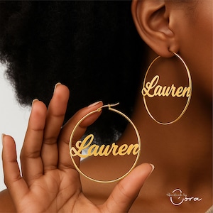 May include: Gold-colored hoop earrings with the name "Lauren" in a cursive font. One earring is held in a hand, and another is worn on an ear. The earrings are a simple, elegant design.