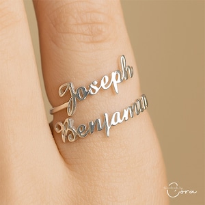 Op de afbeelding: Een zilverkleurige ring met twee banden, elk met een naam in elegant cursief schrift. De bovenste band leest "Joseph", en de onderste band leest "Benjamin". De ring wordt op een vinger getoond, wat het gepersonaliseerde ontwerp benadrukt.