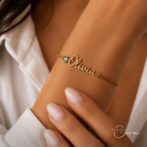 Op de afbeelding: Een goudkleurige kettingarmband met de naam Olivia in een cursief lettertype. De armband bevat een enkele kleine, ronde, lichtblauwe edelsteen in een goudkleurige zetting, geplaatst naast het naamplaatje aan de fijne ketting.