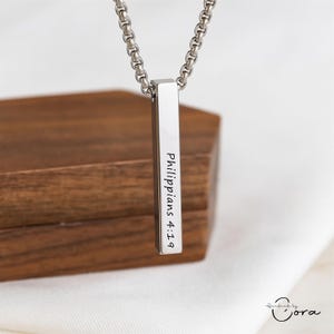 May include: A silver bar pendant necklace inscribed with "Philippians 4:19". The pendant is suspended from a silver chain. The background features a wooden box and a white cloth.