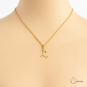 Peut inclure: Collier doré avec un pendentif en forme de lettre "R" orné d'un petit cœur. Le pendentif est suspendu à une chaîne délicate. Le collier est présenté sur une personne.