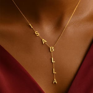 Könnte beinhalten: Eine goldfarbene Halskette mit dem Namen "ISABELLA" in einzelnen Großbuchstaben. Die Buchstaben sind durch eine filigrane Kette verbunden. Die Halskette wird vor einem dunklen Hintergrund präsentiert.