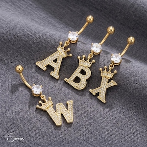May include: Gold-toned belly button rings featuring the letters A, B, X, and W, each adorned with a crown and encrusted with small, clear gemstones. The curved barbell design includes a larger, round, clear gemstone at the bottom.