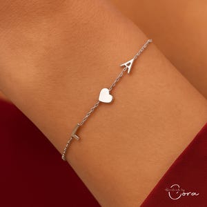 May include: A silver bracelet with the letters "L" and "A" and a heart charm. The bracelet is made of a fine chain and is shown on an arm. The bracelet is a piece of jewellery. The bracelet is a piece of jewellery.