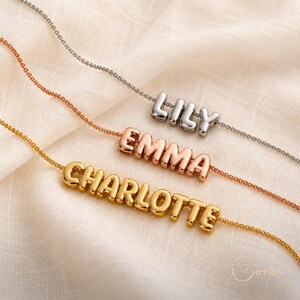 May include: Three delicate chain necklaces featuring polished, rounded, three-dimensional letter charms. The top necklace is silver-toned with the name LILY, the middle is rose gold-toned with EMMA, and the bottom is gold-toned with CHARLOTTE.