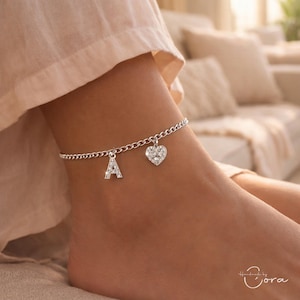 May include: A silver anklet with a chain design, featuring a sparkling letter "A" charm and a heart-shaped charm. The anklet is worn on an ankle, with a soft, neutral-toned background.