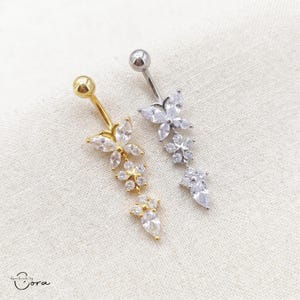 May include: Two belly button rings. One is gold-toned, the other silver-toned, both with a dangling floral design of clear crystals. The gold ring has a gold ball top, the silver ring a silver ball top. The design is reminiscent of flowers.