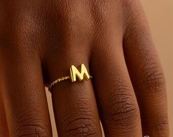 May include: A gold-colored ring featuring the letter "M" in a bold, block style. The ring band has a twisted rope design. The ring is shown on a hand with a warm skin tone.