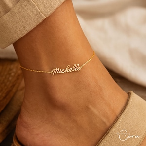 May include: A delicate gold-coloured anklet with the name "Michelle" in cursive script. The anklet is worn on an ankle, with the chain resting just above the foot. The jewellery is a simple, elegant accessory.