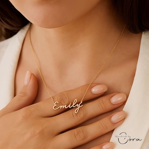 Puede incluir: Un collar dorado con el nombre "Emily" en cursiva. El collar se lleva alrededor del cuello. La persona lleva una blusa blanca. Una joya delicada y elegante.