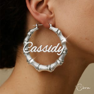 May include: Silver-toned bamboo-style hoop earrings with the name "Cassidy" in a cursive font. The earrings are worn on a person's ear, showcasing the personalized design and the intricate bamboo texture. The earrings are a statement piece.