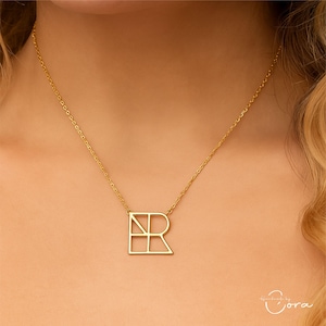Op de afbeelding: Een goudkleurige ketting met een geometrische letter "R" hanger. De hanger is een gestileerd ontwerp met rechte lijnen en hoeken. De ketting heeft een fijne ketting.