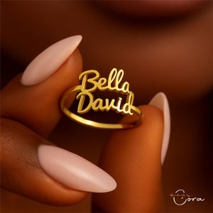 May include: A gold-colored ring with the names "Bella" and "David" in a cursive font. The ring is held between two fingers with manicured nails. The ring is a personalized jewelry item.