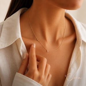 Può includere: Una delicata collana a catena color oro con due piccoli ciondoli a forma di lettera maiuscola, E e A, posizionati in modo asimmetrico lungo il sottile filo metallico. La collana poggia sulla clavicola, mostrando un design semplice.